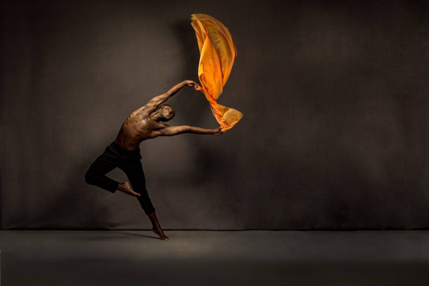 Caption: South Chicago Dance Theatre. Photo credit: Thomas Mohr Photography. Description: ​Barefooted male dancer with a muscular physique and fitted black pants against a dark brown backdrop. His body is facing front, but his head and outstretched arms arc dramatically to the right. His lower torso, hips, and right leg above the knee arc to the left. He is grasping a bright orange scarf that appears to be in mid-air.